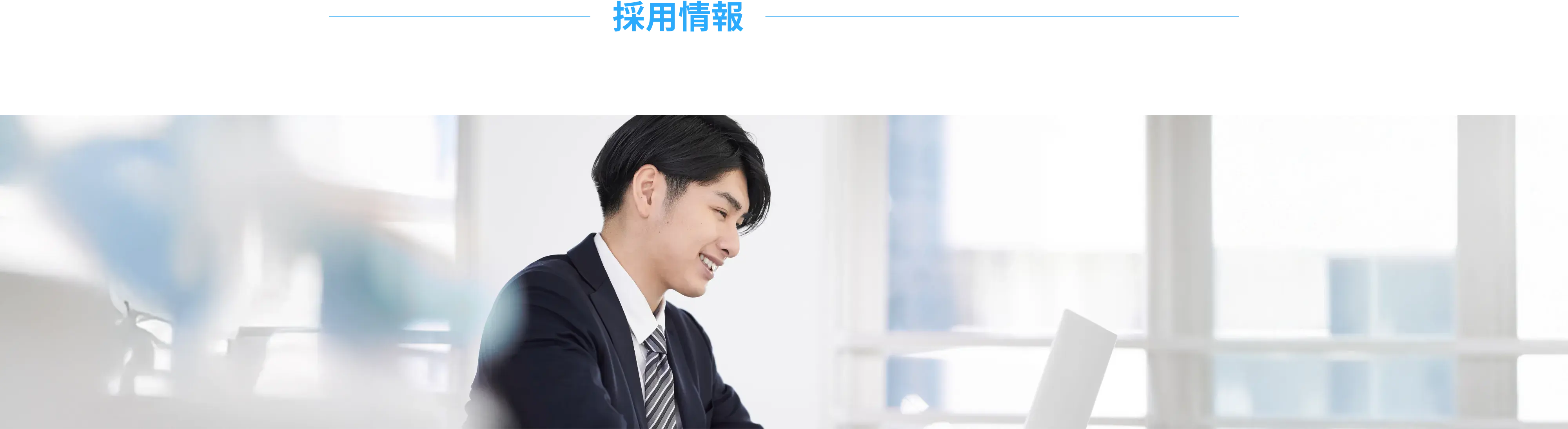 スーツ姿の若い男性がパソコン画面を見ながら微笑んでいるオフィス風景